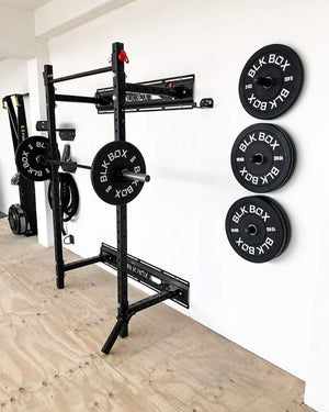A black pull-up bar and squat rack system mounted on a white wall in a home gym setting, with weight plates stacked on the wall and additional equipment visible in the background.