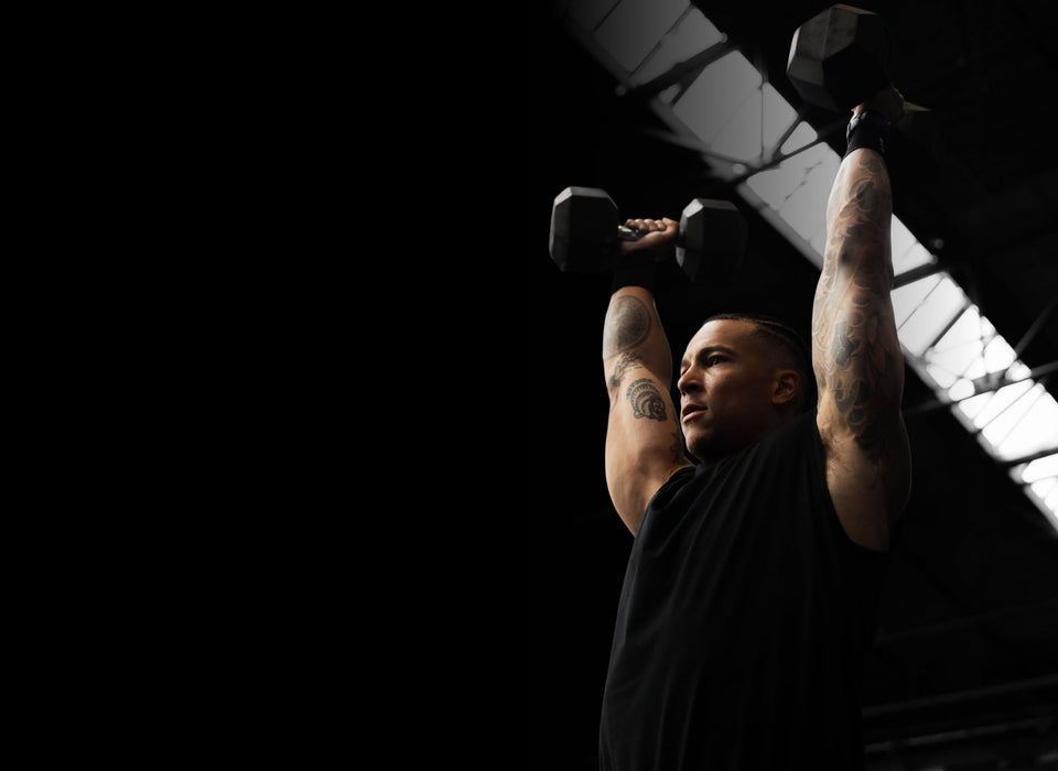 Athlete pressing two hex dumbbells overhead in a dark gym